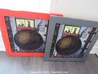 Two framed square paintings leaning against wall, red frame on left and gray on right, both depicting a wrecking ball motif