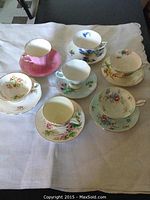 Full view of seven tea cup and saucer sets arranged on white cloth, showing variety in floral patterns, colors, and gold edged rims