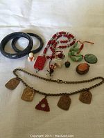 Photo showing all 10 pieces of vintage jewelry spread on a cloth. Two dark blue jade-like bangles, red bead necklace with carved pendant, green bead necklace, small carved charms and pendants, chain with brass charms.