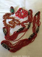 Overall view of five pieces arranged on fabric: two red seed-bead necklaces, one amber-tone bead necklace, one red coral-chip bracelet, one silver-tone ring.
