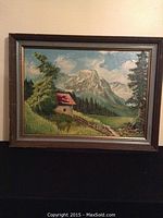 Framed oil painting showing a mountain landscape with a cottage, trees, and sky.