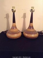 Pair of two Mid Century ceramic table lamps with bulbous lava glazed bases and tapered necks, sitting side by side on black surface against beige wall.