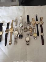 16 assorted wristwatches laid out on white fabric, varying metal link and leather strap types