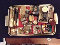 Photo showing a tray full of assorted small collectibles including dice, pins, badges, mini figurines, keychains, lighters, and a small metal spoon.