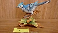 Porcelain blue jay figurine perched on a branch with leaves and acorns in front of wooden backdrop.