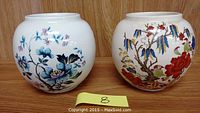 Two ceramic ginger jars shown side by side against a wooden background. One jar features a blue floral design, the other a botanical red and blue floral design.