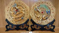 Two decorative gold filigree plates with intricate metalwork and colorful enamel details mounted on black stands.