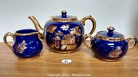 Teapot, creamer, and sugar bowl arranged side by side showing gold handles and blue glaze with gold Asian-style artwork.