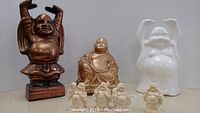 Photo showing three larger Buddha figurines including a raised-arm bronze figure, a gold-painted seated Buddha, and a glossy white ceramic raised-arm Buddha, along with the five smaller carved cream-colored Buddhas.