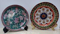 Two decorative plates displayed side by side on stands: one with green floral design and one with multicolored floral-geometric pattern