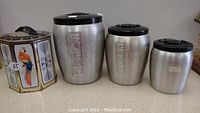 Set of four kitchen storage containers displayed on a counter, including the hexagonal container and three cylindrical metal containers with black lids.
