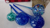Four glass globes laid side by side, various shades of blues and green with long tapered necks and round reservoirs.