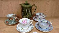 Group photo showing four assorted tea cups with matching saucers alongside a green ceramic tea pot with textured decorative pattern.
