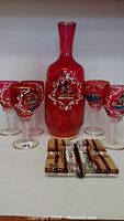 Full view of red Murano glass decanter with four matching wine glasses, all decorated with white and gold Italian landmark motifs.