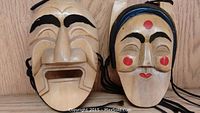Photo shows two wooden masks, one with an open mouth and prominent eyebrows, the other with painted black hair and red markings on face.