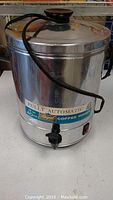 Photo showing full view of stainless steel coffee maker with lid, power cord, and side handle.