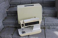 Singer Memory Touch Tronic 2010 zig-zag sewing machine on stone steps with accompanying beige carrying case behind it.