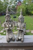 Two silver gilt kneeling figurines shown outdoors on wooden surface with greenery background. Both have intricate headgear and traditional attire, hands in prayer gesture.
