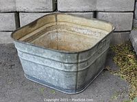 Square galvanized metal wash tub sitting on pavement, showing weathered exterior with some rust and wear.
