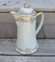 Front view of an antique porcelain chocolate pot with gilded accents and hand-painted floral designs, photographed on wooden steps.