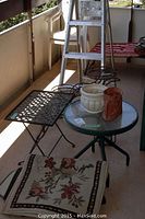 Metal ladder, small round table with glass top and metal base, two planters, and rugs on balcony floor.