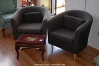 Two grey tub-style arm chairs with cylindrical cushions and an antique wooden foot stool with floral fabric cushion arranged on wooden floor.
