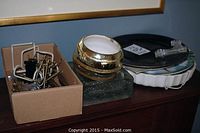 Assorted collector plates with some decorative borders, a box of plate hangers, and two distinct glass items on a wooden surface.