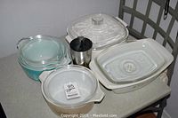 Photo showing six casserole dishes with lids and a metal cup on a table.