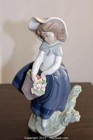 Front angled view of Lladro figurine showing girl with basket of flowers, wearing blue dress and hat.