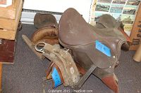 Two leather English saddles stacked on carpet floor, one labeled 22X16 and the other 17X19.