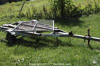 Side angle view of Highlander utility trailer on grass, showing metal frame, wooden bed support beams, and mesh fold-down gate on trailer bed.