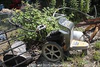 Side view of the Club Cadet mower showing wheels, handle and green body, in an outdoor environment with vegetation around it.