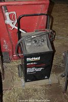 DieHard battery tester with controls and attached cables in front of large red gas container with wheels and fuel nozzle.