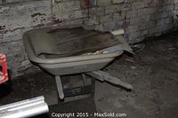 Side view of large wheelbarrow with basin filled with rolled mats and a wooden dowel, showing wear and stored indoors.