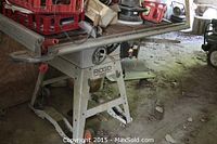 Side view of Rigid brand table saw with extendable table and frame with wheels, placed on a dusty floor with other tools in background.