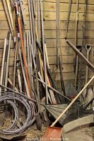 Wide view showing multiple wooden-handled tools including rakes, shovels, a coiled roll of wire, and metal tools leaning against a barn wall with hay on the floor.
