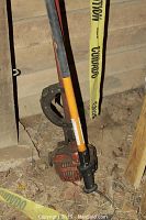 Long gas-powered pole pruner saw leaning against a wall, showing orange handle, black pole, and engine base.