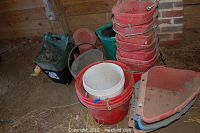 Assortment of plastic buckets shown stacked on barn floor, including red, white, green, and black buckets