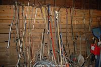 Wide angle photo showing multiple gardening tools on the back wall including rakes, shovels, scythes, and other hand tools with wooden handles and metal parts.