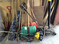 Wide view of assorted long-handled tools, spreader, edger attachment, gas engine and hand tools leaning against wall