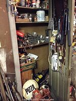 Closet interior with shelves containing assorted paint cans, spray cans, various hardware boxes and parts, and a white pizza sign leaning against lower shelves.