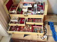 Cream wooden jewelry box opened showing three drawers with red felt-lined compartments filled with jewelry