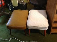 Pair of vintage hassocks side by side on a green carpet floor, showing cushions and legs.