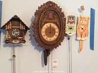 Four clocks mounted on wall: wooden chalet cuckoo clock, carved wooden wall clock with Spanish text, cat-themed calendar clock, and cat-shaped clock with pendulum tail.