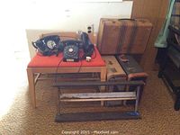 Two Bakelite rotary dial telephones placed on an orange-cushioned wooden chair, with three vintage suitcases in the background and a paper cutter in front.