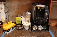 Full view of Bosch Tassimo coffee maker with wire rack holding sealed coffee pods and coffee accessories including reusable filter and a sugar bowl.