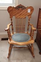 Full view of the wood rocking chair showing carved headpiece, armrests, and turned wood spindles with cushion on seat.