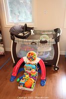 Brown plaid Graco playpen with mesh siding and infant seat with white plush toy inside, accompanied by assorted colorful baby toys on the floor in front.