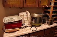 Wide shot of lot on kitchen counter including red enamel electric roasting pan with lid, white Moulinex deep fryer, Presto kitchen kettle, and large stainless steel pot.