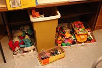 Wide view of three boxes with children's toys under a desk. Left box contains plush Smurf dolls, middle is a yellow trash bin, right boxes contain toy vehicles and parts.